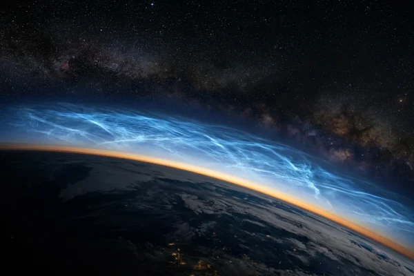Electric blue noctilucent clouds glowing above a dark horizon against a starry night sky.