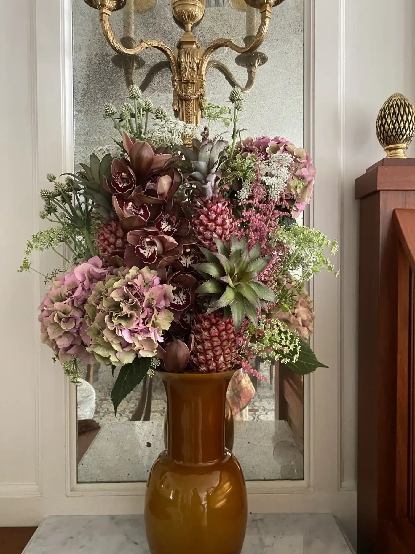 A floral arrangement in a vase with a decorative mirror and ornate fixture in the background