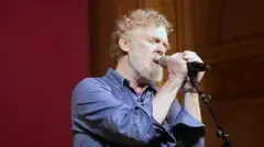 Getty Images Glen Hansard has short curly reddish/brown/grey hair and a greyr beard and is singing with two hands on the microphone and his eyes closed
