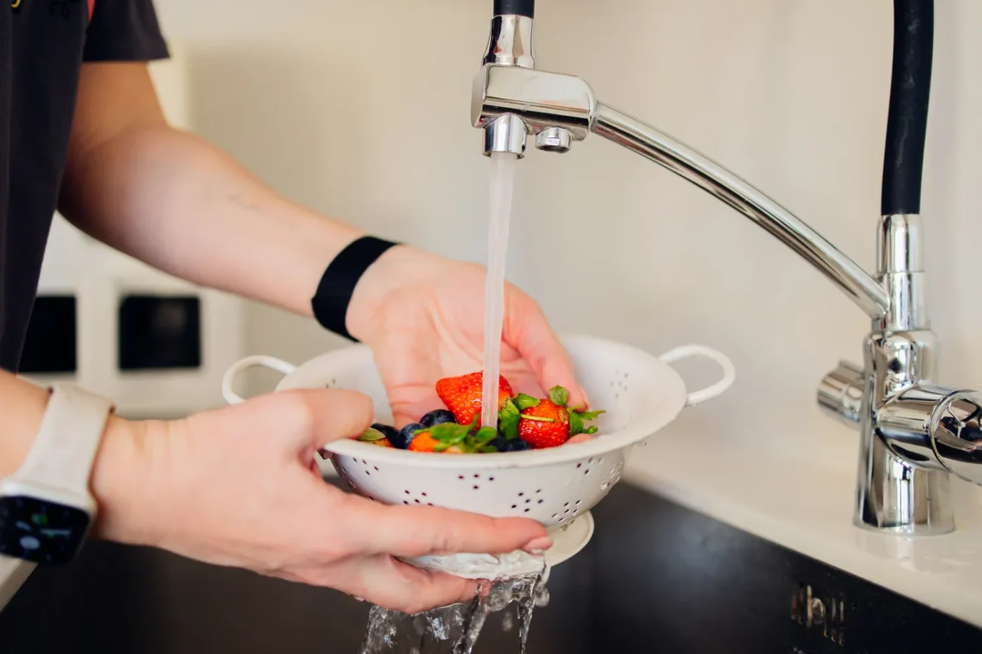 Hands washing fresh produce in a colander under a running faucet