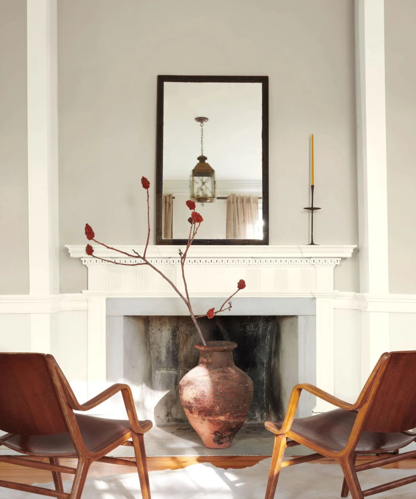 Living room with large white fireplace with a rustic pot holding foliage, walls painted in Benjamin Moore's Revere Pewter, a large mirror on the mantelpiece, and two midcentury leather lounge chairs.