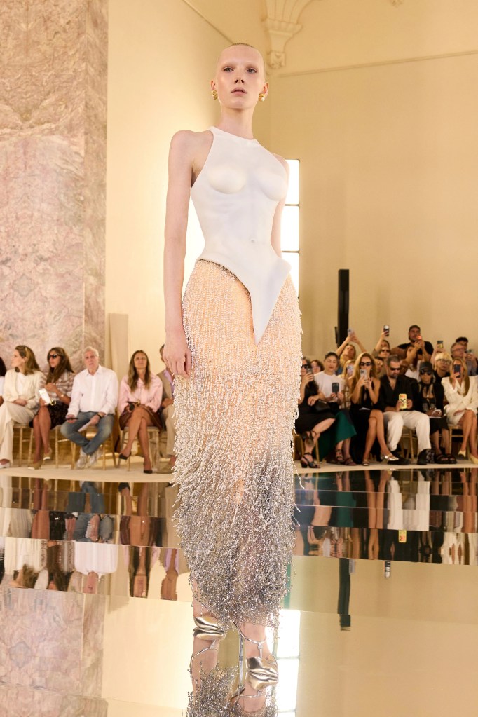 A model on a runway wearing a white sculpted bodice and a long, beaded skirt that fades from peach to silver.