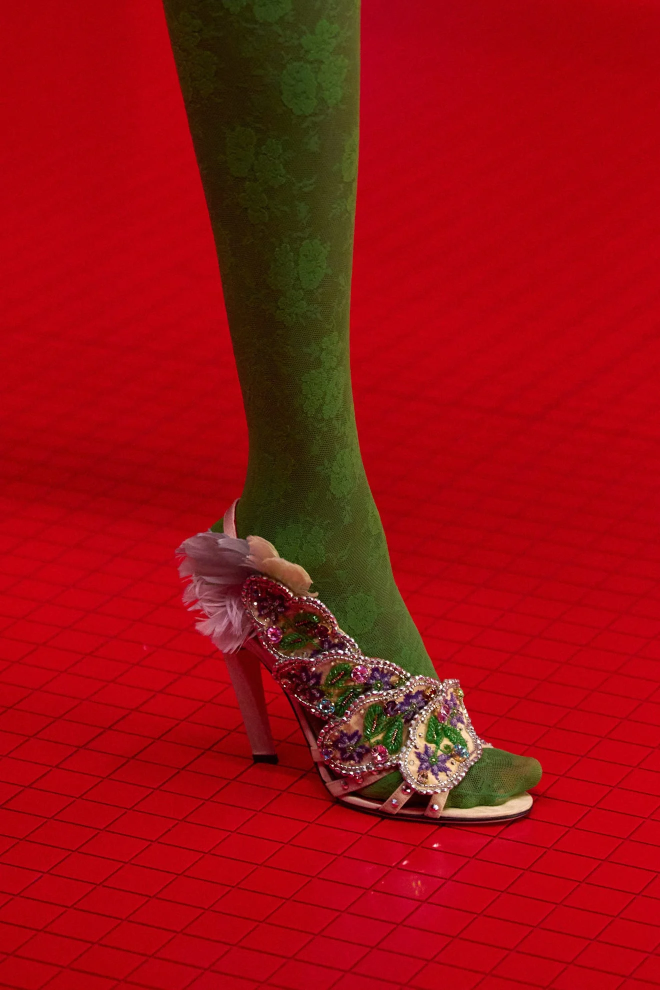 Decorative high-heeled shoe with embellishments, worn with green patterned tights on a red textured surface.