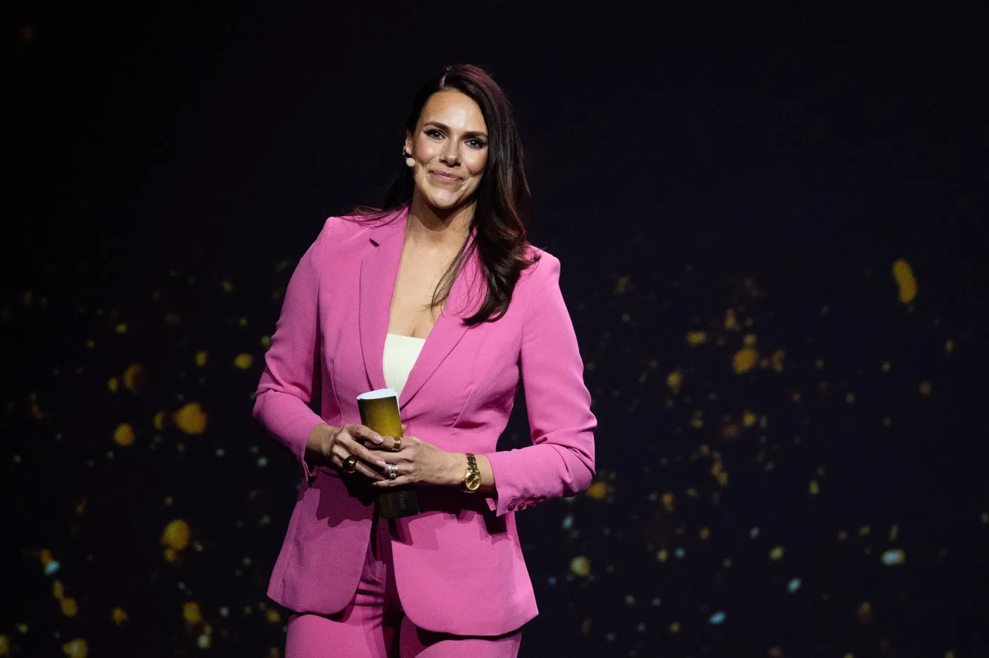 In einem pink-elegantem Anzug moderiert Esther Sedlaczek mit gefalteten Moderationskarten in den Händen eine Sportgala.