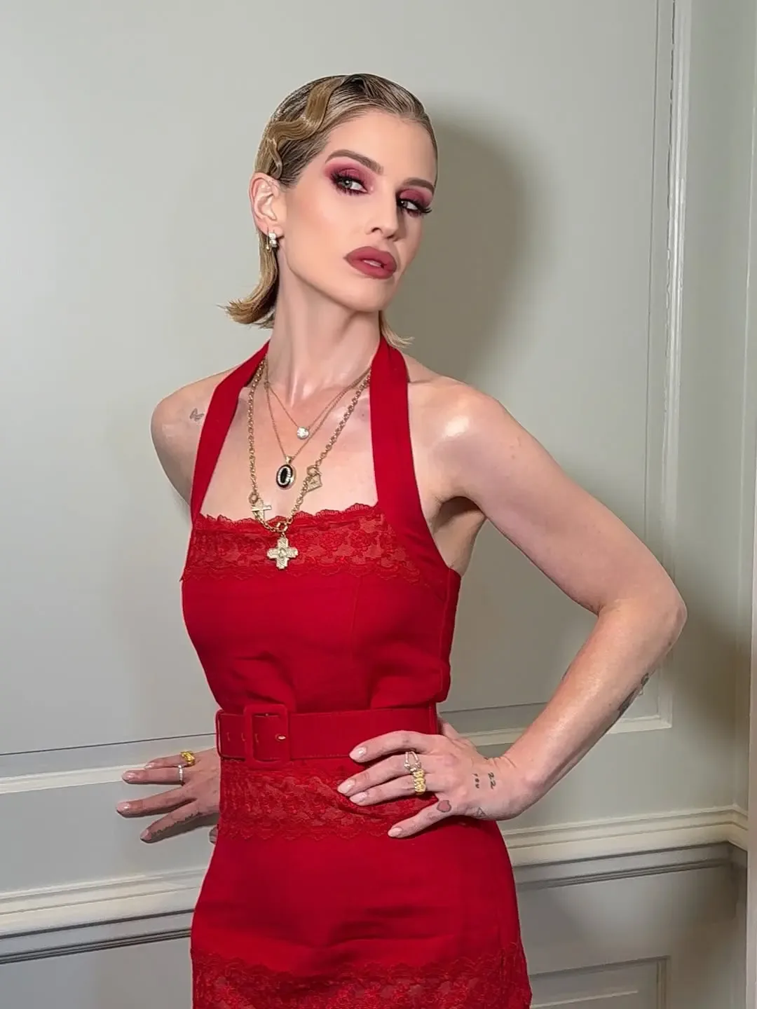 Kelly Osbourne in a red halter-neck dress with lace trim and gold necklaces, hands on hips.