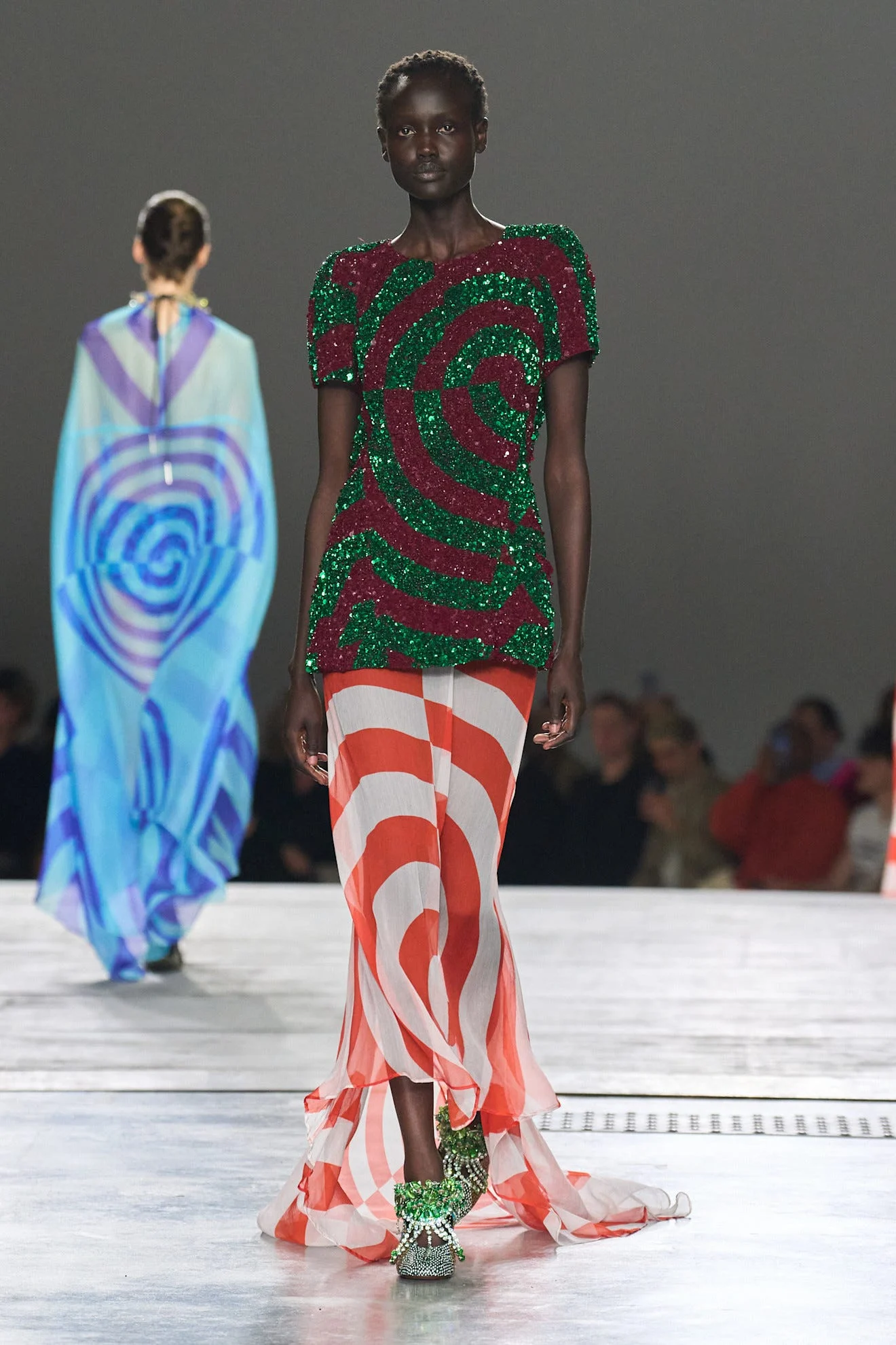 Model walking the runway in a sparkling green and red spiral top with sheer orange and white striped skirt and embellished shoes.